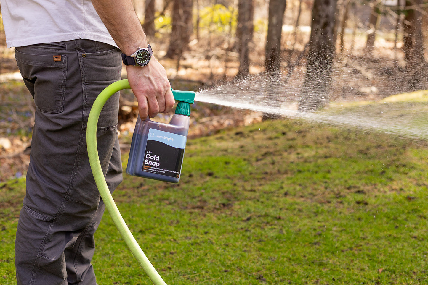 Person using a spray bottle labeled 'Cold Snap' to water grass outdoors.